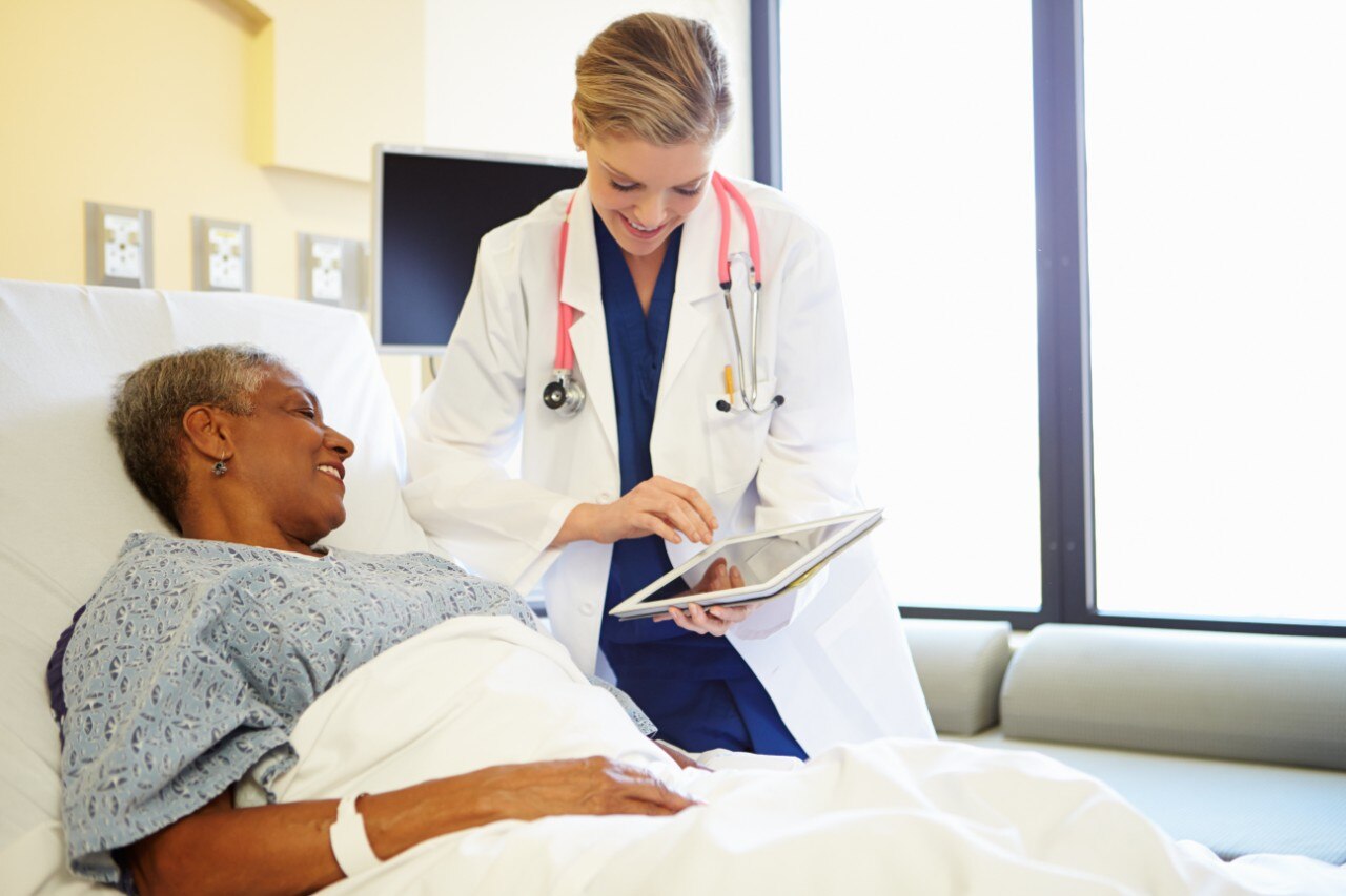 Medical professional helping a patient by showing her something on the tablet with patient in bed.