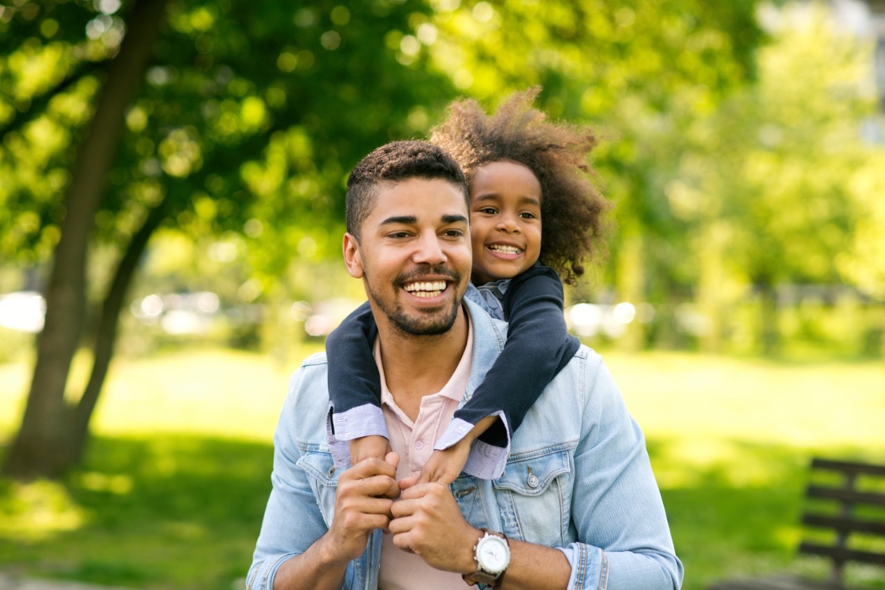 A man and his child take a walk in the park on a beautiful day with him giving her a piggyback