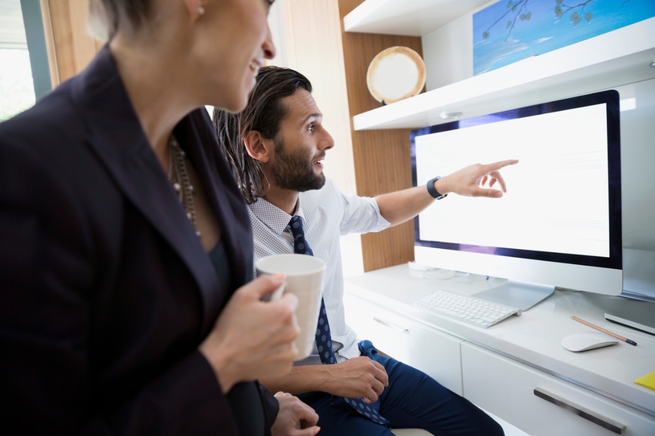 Male business professional pointing to the screen of his big computer monitor showing another business professional something on the screen.