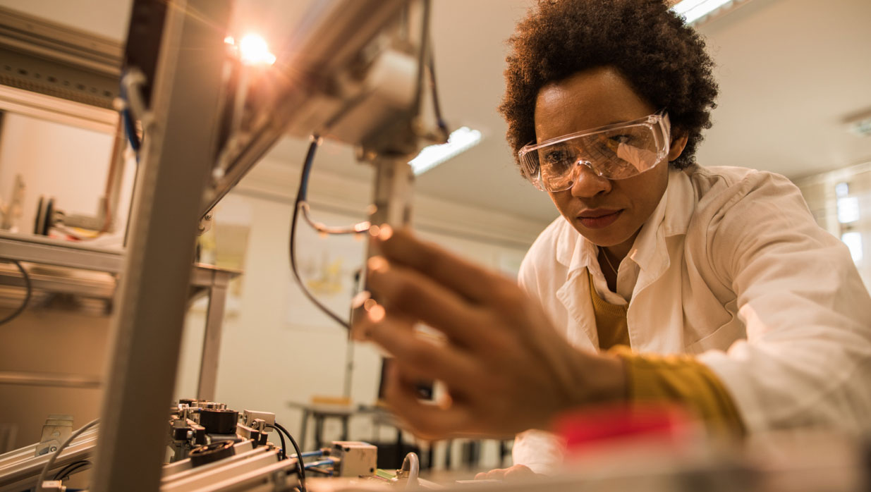 Female lab technician in safety goggles & lab coat inspecting product components