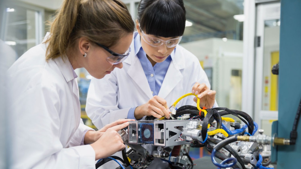 Two female lab technicians in protective gear at manufacturing plant with diagnostic equipment testing product components